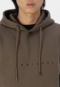 Brun hættegenser med snore og sort logo tekst "JACK & JONES" på fronten. Lavet af blødt materiale med en glat tekstur.