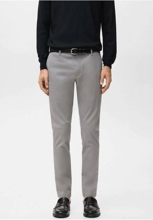 Mann trägt schmal geschnittene hellgraue Hose, schwarzen Gürtel, schwarzen Langarmoberteil und schwarze Lederloafer, steht vor einem einfachen Hintergrund.