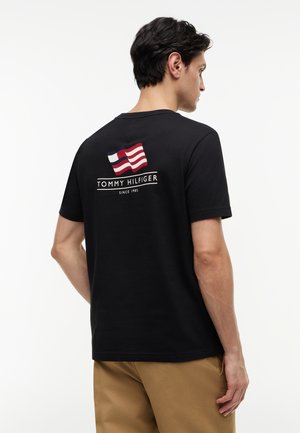 Hombre de espaldas vistiendo una camiseta negra de Tommy Hilfiger con una bandera en rojo, blanco y azul y el texto "TOMMY HILFIGER DESDE 1985" en la parte trasera.