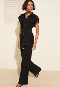 Zwarte jumpsuit met korte mouwen, knoopsluiting aan de voorkant, ceintuur in de taille, twee borstzakken en rechte, brede pijpen. Glad materiaal en minimale details.