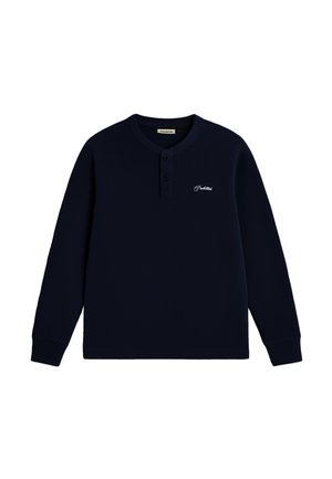 Jeune homme portant une casquette bleu marine et une chemise henley à manches longues assortie avec un logo brodé, debout, une main dans la poche.