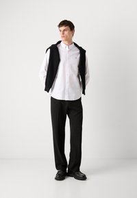 Λευκό πουκάμισο με κουμπιά και μαύρο πουλόβερ ριγμένο στους ώμους, μαύρα παντελόνια και μαύρα παπούτσια, σε ανοιχτό γκρίζο φόντο.