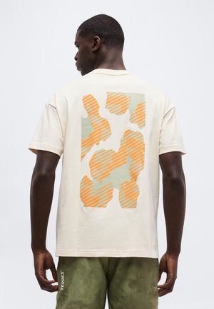 Homme portant un t-shirt crème avec des formes abstraites orange et vert clair dans le dos, et un short camouflage vert avec le texte « TERREX ».