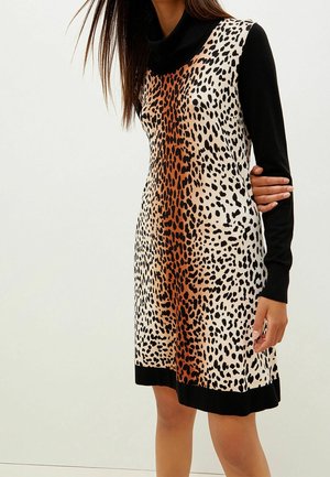 Kobieta w sukience bez rękawów w panterkę, z czarnym golfem i długimi rękawami, stoi na tle jasnego, jednolitego tła.