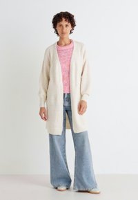 Lång cardigan med öppen front i mjuk beige. Har vida ärmar och är matchad med en rosa texturerad tröja och ljusblå utsvängda jeans.