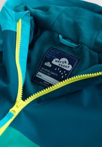 Chaqueta impermeable color verde azulado con cremallera amarilla, forro polar y etiqueta en el interior del cuello que dice "Impermeable, La aventura espera, Hecho en Bangladesh".