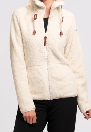 Femme portant une veste polaire blanche à fermeture éclair avec cordons et accents en cuir, associée à un pantalon noir.