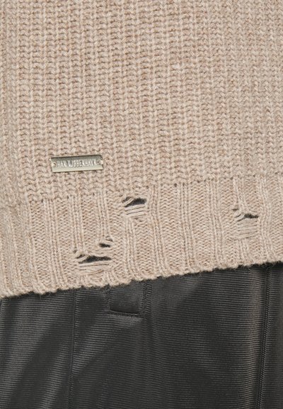 Μπεζ πλεκτό πουλόβερ με ριγέ στρίφωμα, που διαθέτει μικρές φθαρμένες τρύπες και μεταλλική πλάκα με λογότυπο. Φορεμένο πάνω από μαύρα υφασμάτινα παντελόνια.