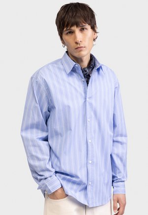 Νέος άνδρας με κοντά καστανά μαλλιά, φορώντας γαλάζιο πουκάμισο με κάθετες ρίγες, σκούρο φουλάρι με σχέδιο στον λαιμό και λευκό παντελόνι, στέκεται με το ένα χέρι στην τσέπη.