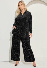Set di pigiama in velluto nero composto da una camicia con bottoni e colletto e pantaloni larghi, entrambi decorati con pois bianchi a forma di stella sparsi.