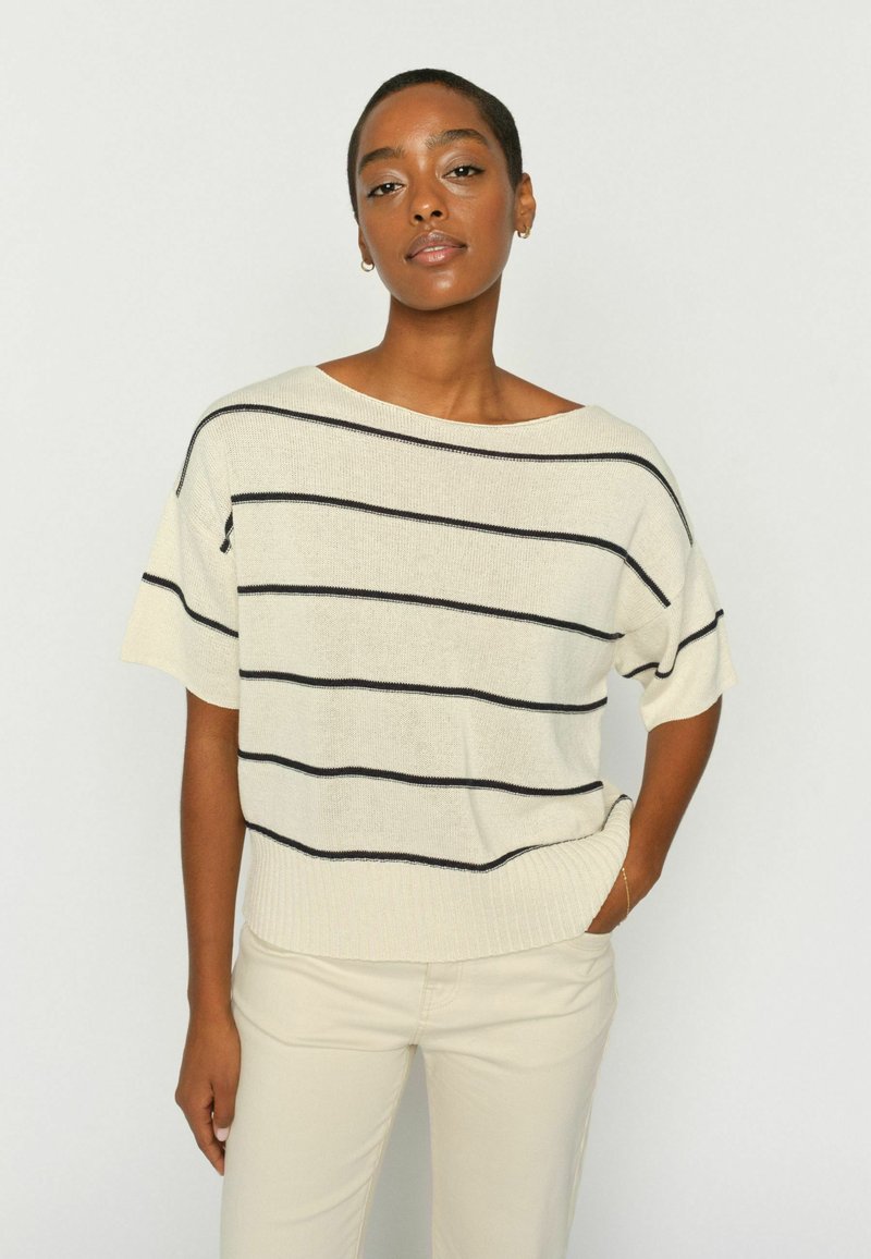 Žena u kremasto bijelom džemperu s kratkim rukavima s crnim vodoravnim prugama i svijetlo bež hlačama, stoji s jednom rukom u džepu.