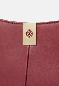 Detailní záběr na burgundskou koženou kabelku s obdélníkovým zlatým zapínáním, které má vyřezávaný kruhový květinový vzor.