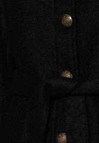 Svart yllekappa med texturerad finish, bronsknappar med detaljerad design och ett tygskärp för justerbar stängning.