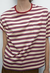 Henkilö, joka käyttää lyhythihaista kermaan vivahtavaa T-paitaa, jossa on punaisia ja sinisiä vaakasuoria raitoja ja punareunainen pyöreä pääntie valkoista taustaa vasten.