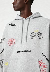 Felpa grigia realizzata in morbido tessuto, con vari design ricamati: una rosa rossa, ali d'angelo nere e gialle e incisi di testo in nero.