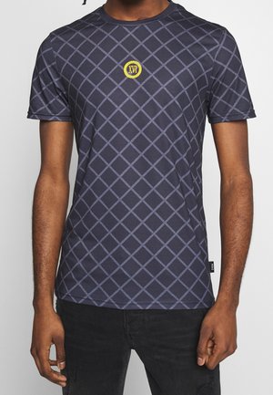 Man wearing navy blue T-shirt with light gray diamond grid pattern and small yellow circular emblem with "XVI" on chest.
