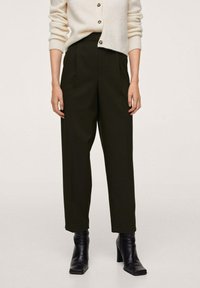 Donkergroene, hooggeremde broek met plooien; gecombineerd met een crèmekleurig vest met knopen, gedragen met zwarte enkellaarsjes met hak.