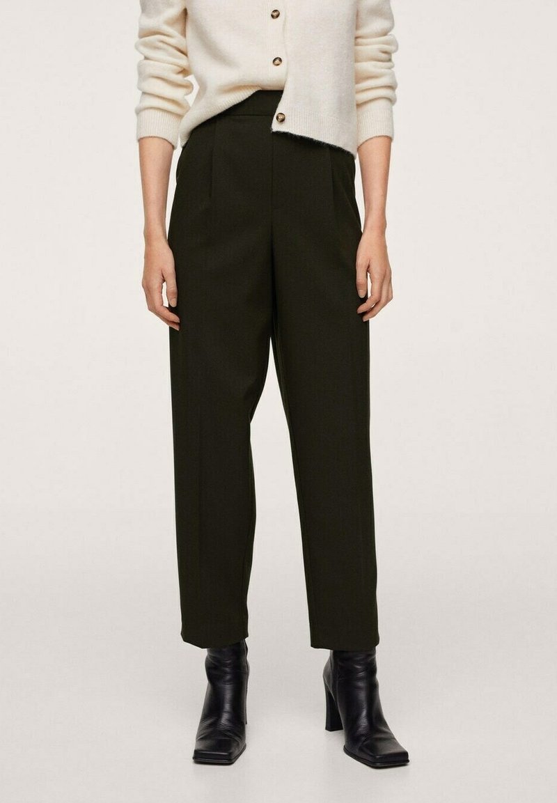 Donkergroene, hooggeremde broek met plooien; gecombineerd met een crèmekleurig vest met knopen, gedragen met zwarte enkellaarsjes met hak.