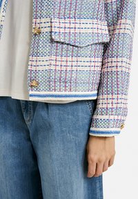 Gewebte Jacke in Pastellfarben mit Karomuster, goldenen Knopfakzenten und blauen Akzenten an den Ärmeln, kombiniert mit hellblauen Jeans.