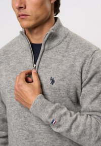 Pilkas mezgimo megztinis su aukštu apykakle ir užtrauktuku, su mėlynu polo logotipu ant krūtinės ir trikampiu akcentu ant rankogalių.