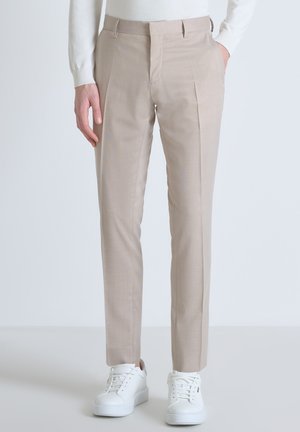 Pantaloni beige su misura con una texture liscia, caratterizzati da una parte anteriore piatta, tasche laterali e passanti per cintura, abbinati a sneakers bianche.