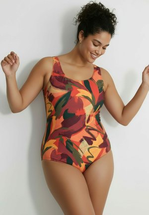 Kobieta o ciemnych kręconych włosach, ubrana w kolorowy kostium kąpielowy jednoczęściowy z abstrakcyjnym wzorem, uśmiechająca się i unosząca obie ręce na białym tle.