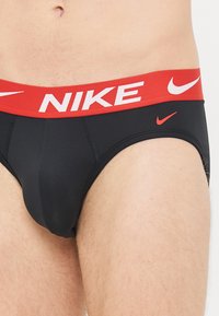 Black men's briefs with red elastic waistband featuring white "NIKE" text. Smooth fabric, fitted design, and small red logo accent.