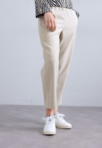 Pantaloni beige su misura con una texture liscia, caratterizzati da una vita media e gambe affusolate, abbinati a sneakers bianche.