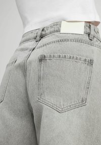 Jeans en denim gris avec une taille haute, comportant deux poches arrière et une étiquette en cuir blanche au niveau de la taille, mettant en valeur un tissu texturé.