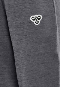 Närbild av grått tyg med ett litet broderat bi-logotyp i vit och svart tråd.