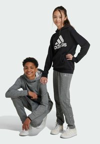 Conjunto deportivo gris con una parte superior con capucha y pantalones de chándal que presentan detalles en negro. La parte superior tiene un logo blanco; ambos llevan zapatillas blancas.