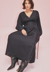 Juoda ilga rankovė su giliu V formos iškirpimu, suspausta juosmeniu ir klostuota sijonu. Priderinta prie taškinių juodų odinių aulinukių iki kulkšnies.