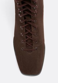 Stivale alla caviglia in suede marrone con punta affilata, dotato di lacci incrociati e occhielli in metallo, che mette in evidenza una texture liscia e finiture lucide.