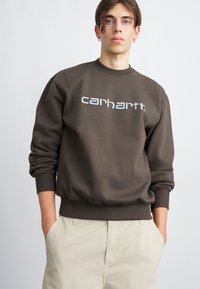 Pulover maro cu manșete și tiv cu nervuri, având logo-ul alb "carhartt" pe piept. Purtați cu pantaloni bej deschis.