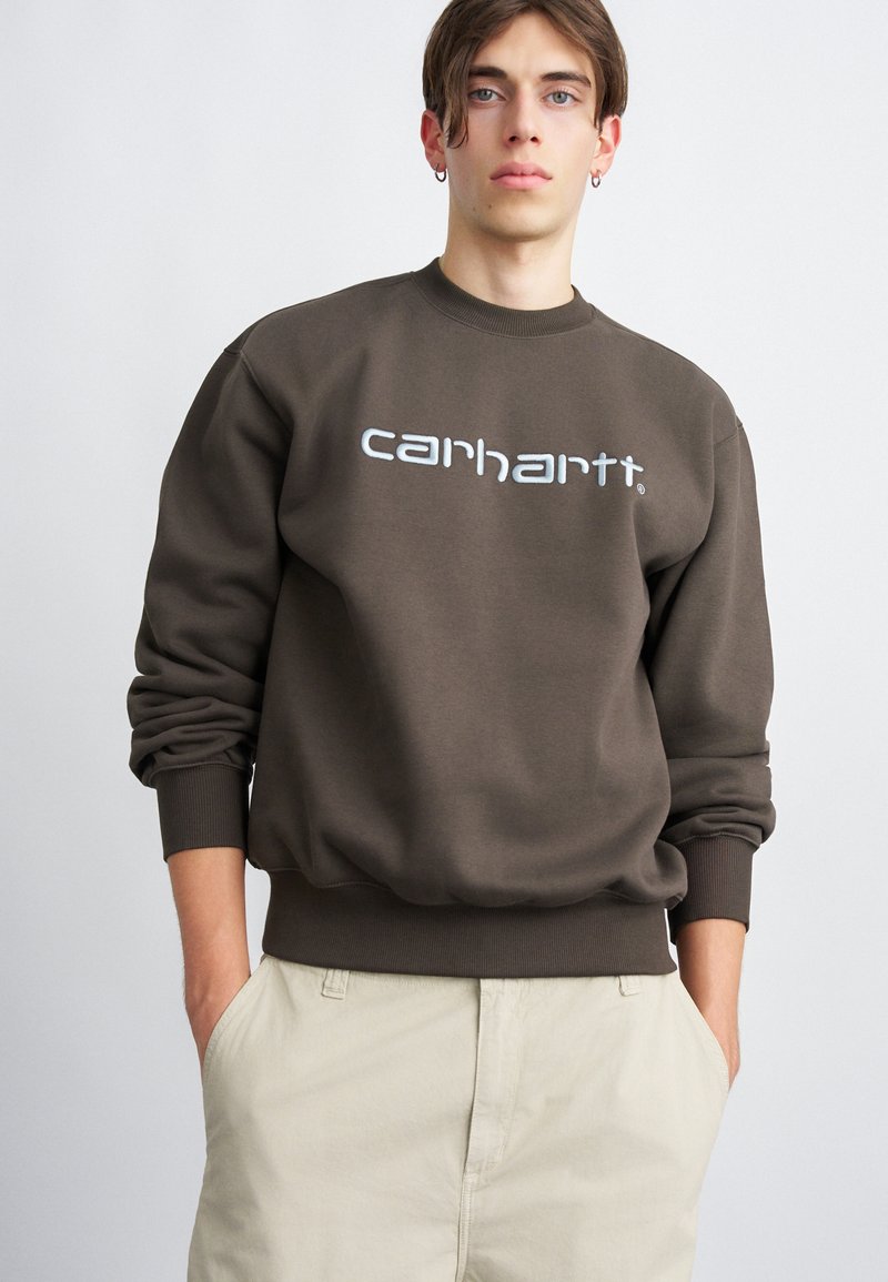 Pulover maro cu manșete și tiv cu nervuri, având logo-ul alb "carhartt" pe piept. Purtați cu pantaloni bej deschis.