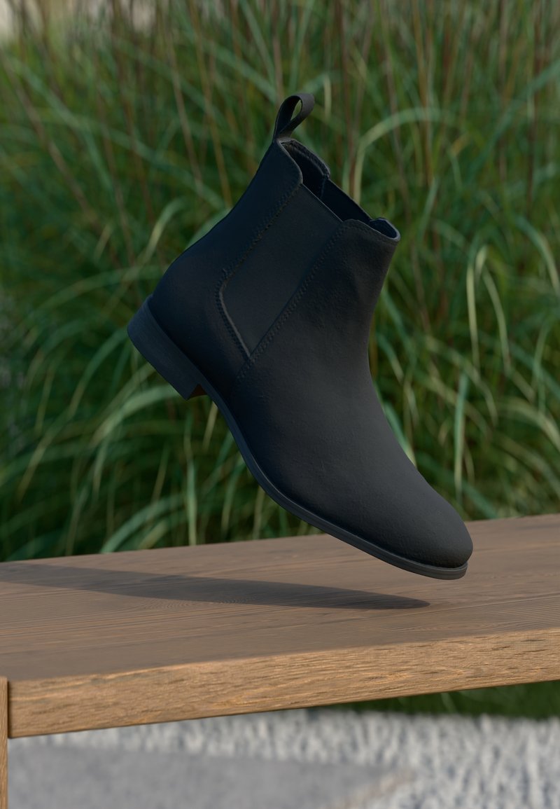 Botte Chelsea noire suspendue au-dessus d'un banc en bois avec de hautes herbes vertes floues en arrière-plan.