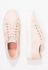 Une paire de baskets basses rose clair avec des semelles blanches, des œillets dorés, des lacets roses et des étiquettes en textile de marque sur la languette et le côté.
