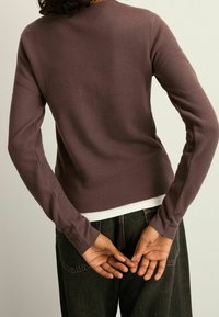 Osoba w ciemnofioletowej bluzce z długim rękawem i czarnych spodniach stoi tyłem, z rękami splecionymi za plecami na jednolitym tle.