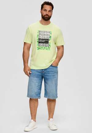 Lichtgroene katoenen t-shirt met een grafische print met verschillende teksten en een rand van groene ballen, gecombineerd met blauwe denim shorts en witte sneakers.
