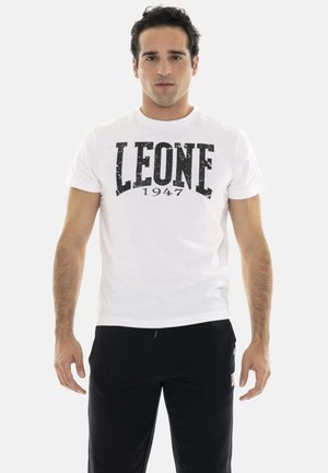 Man die frontaal staat, draagt een wit "LEONE 1947" T-shirt en zwarte broek tegen een effen achtergrond.