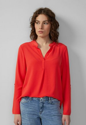 Langsleeve shirt met diepe V-hals in felrood, met een textuurstof en een subtiele naaddetail bij de halslijn. Gematcht met blauwe jeans.