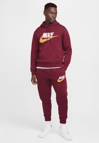 Conjunto de sudadera con capucha y pantalones de chándal en color burdeos, hecho de una mezcla de algodón. Presenta grandes logos de Nike en blanco y naranja. Zapatillas blancas completan el atuendo.