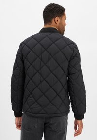 Homme portant une veste matelassée noire avec un motif en losange, se tenant de dos face à l'appareil photo sur un fond uni.