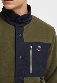 Fleece jacket with olive green body and navy blue collar. Features a zippered chest pocket and snap buttons on the front.