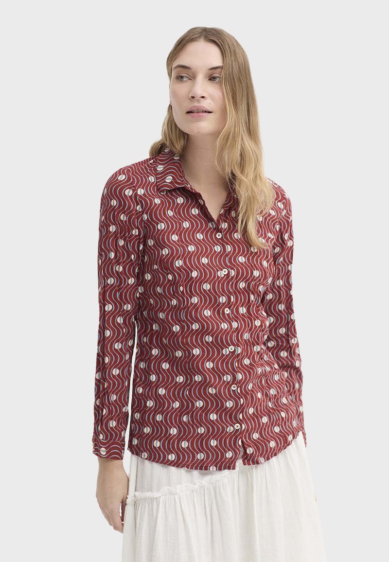 Chemise rouge à manches longues avec un motif ondulé blanc et bleu, col à boutons et texture lisse, portée sur un vêtement blanc texturé.