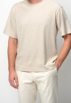 Man wearing a plain beige short-sleeve T-shirt and light-colored pants with one hand in pocket against a plain background.
