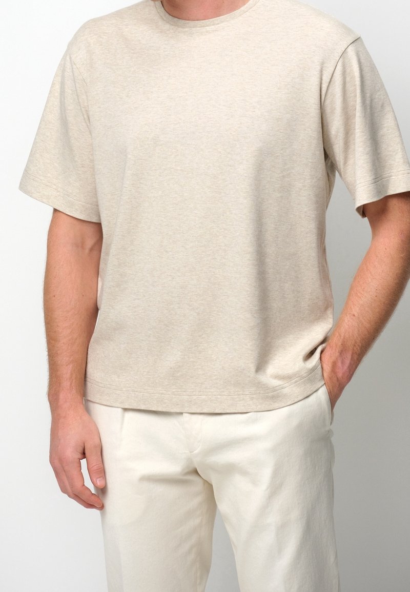 Homme portant un T-shirt beige uni à manches courtes et un pantalon clair, une main dans la poche, sur un fond uni.