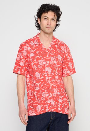 Mann mit dunklem lockigem Haar trägt ein rotes Kurzarmhemd mit weißen Blumen- und Pagodenmustern und steht vor einem schlichten Hintergrund.