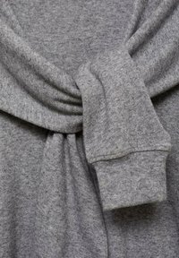 Grauer Strickpullover mit einem geknoteten Vorderteil, weichem Material und angeschnittenen Ärmeln. Sichtbare Nähte umrissen den Stoff.