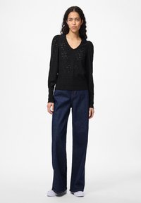 Svart strikkegenser med V-hals og puffede skuldre, kombinert med mørkeblå jeans med vide ben og hvite joggesko.